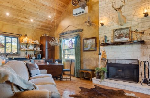 Large great room with upholstered sofa, hardwood flooring, stone fireplace, cowhide rug, kitchen, and dining area