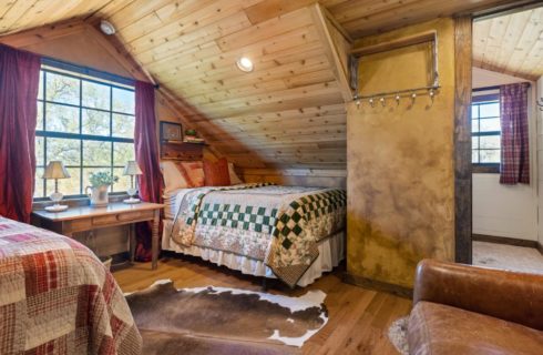 Bedroom with two beds, hardwood flooring, wood paneling on the ceiling, leather sofa, cowhide rug, and view into bathroom
