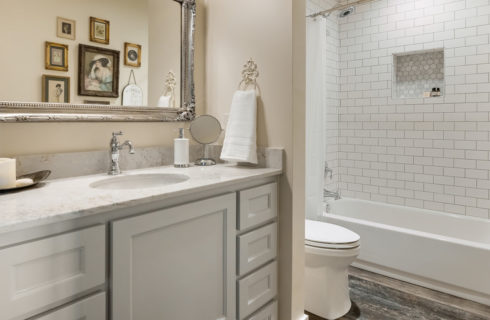 White vanity with many drawers and marble top and silver-framed mirriro above. white tiled shower/bath and toilet beside.