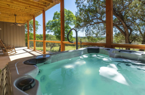 Filled hot tub with cup holders underneath wooden covered porch and rocking chairs surrounded by trees.