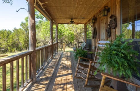 Long wooden covered porch with railing facing woods with rocking chairs on each side of door in front of windows and ferns on stands in between.
