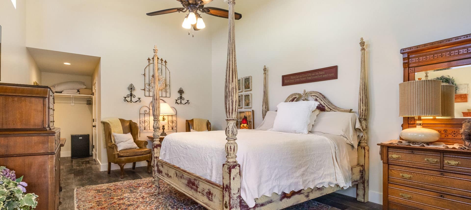 Large bedroom with four-poster bed, white bedding, dark hardwood flooring, large area rug, wooden furniture, and celing fan