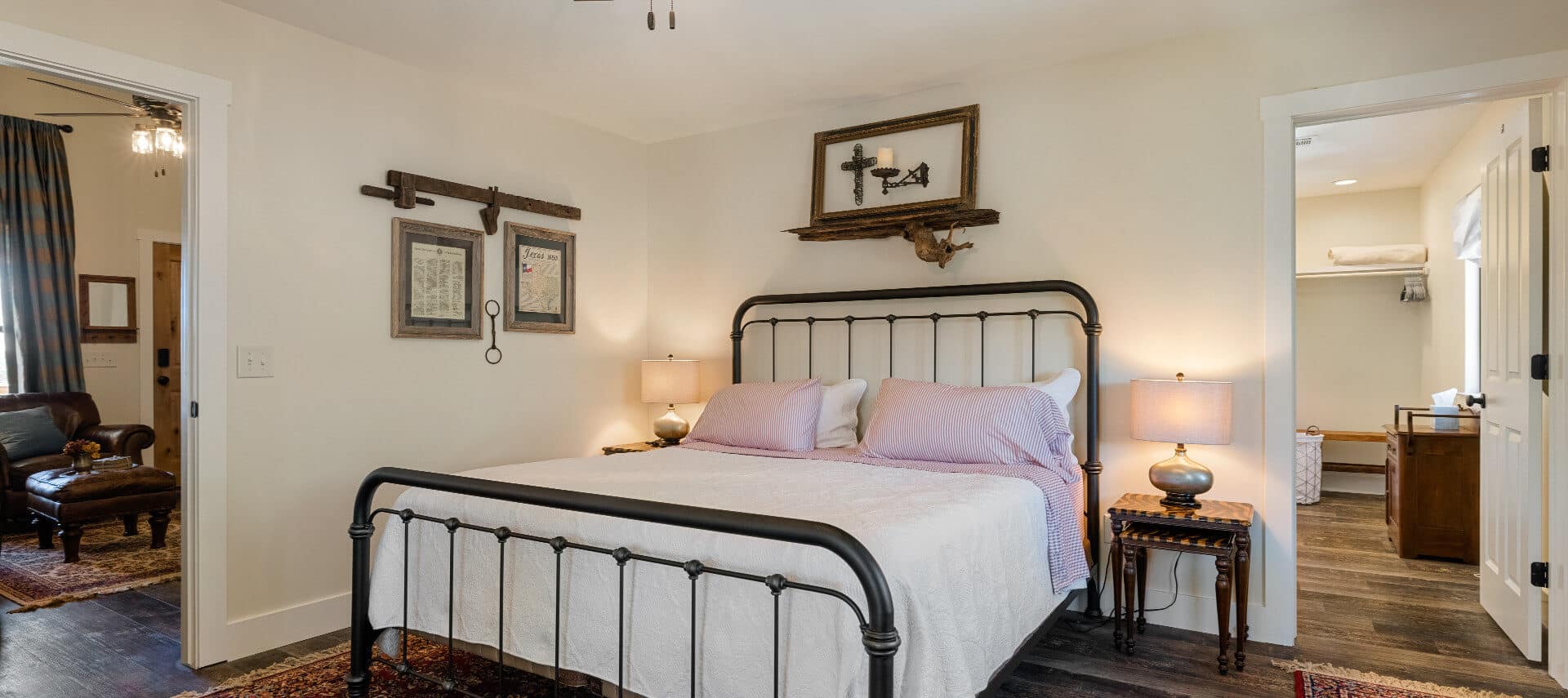 Wrought iron bed in center room with cream walls with framed pictures on 2 walls and nightstand, wood floors with 2 connected rooms on each side.