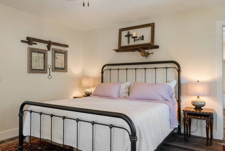 Wrought iron bed in center room with cream walls with framed pictures on 2 walls and nightstand, wood floors with 2 connected rooms on each side.
